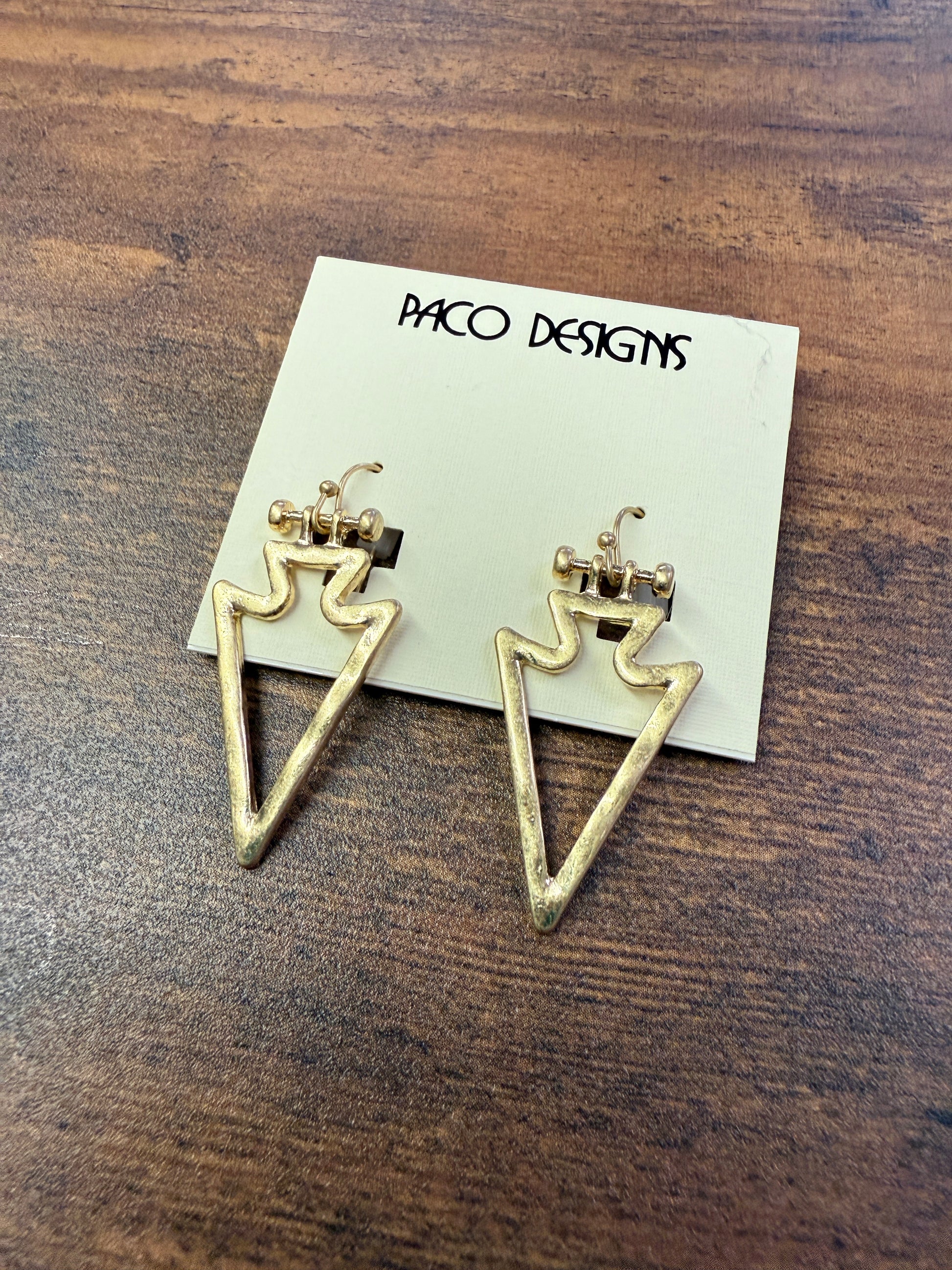 Gold triangle-shaped earrings on a wooden surface with 'Paco Designs' card.