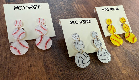 Three pairs of sports-themed earrings on cards with 'Paco Designs' branding on a wooden surface.