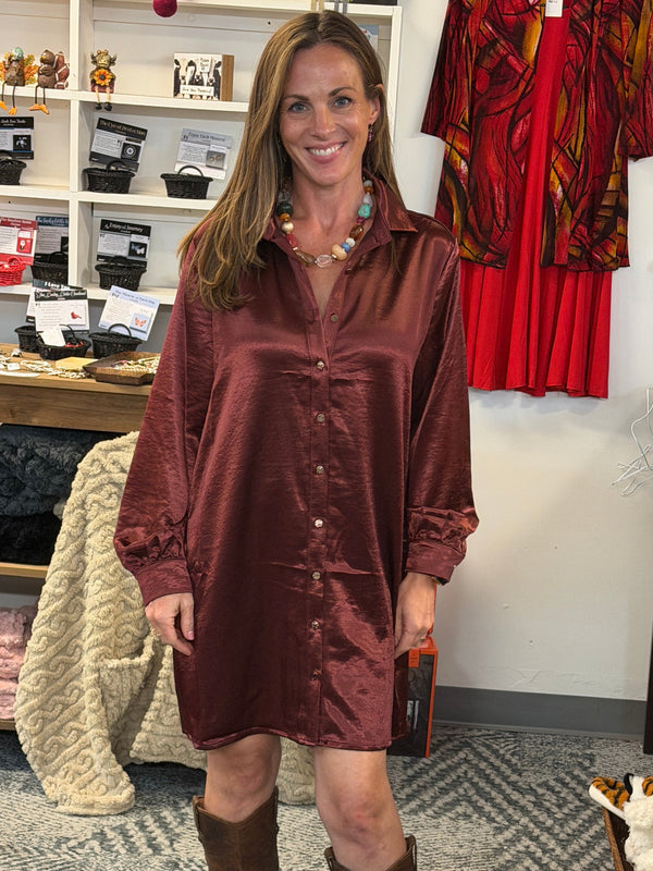 A woman stands in a retail store wearing a shiny burgundy satin shirt dress with brown knee-high boots. The store has shelves with various items, a red patterned jacket hanging on the wall, and a gray patterned carpet.