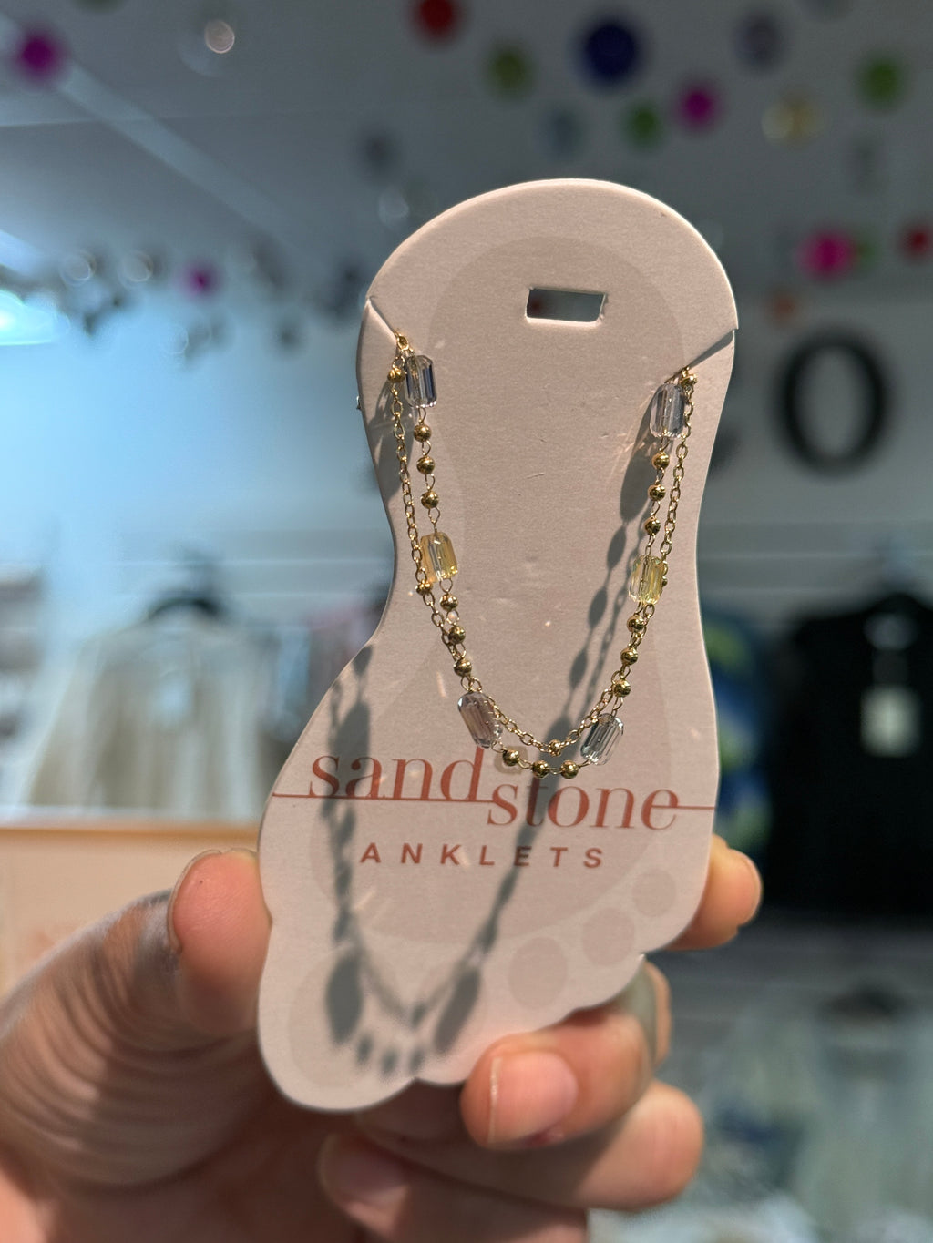 Gold anklet on a 'Sandstone Anklets' card held by a hand with a blurred background