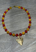 A beaded necklace with alternating translucent red and yellow faceted beads, featuring a hammered gold-tone arrowhead pendant, displayed on a gray wood-textured surface.