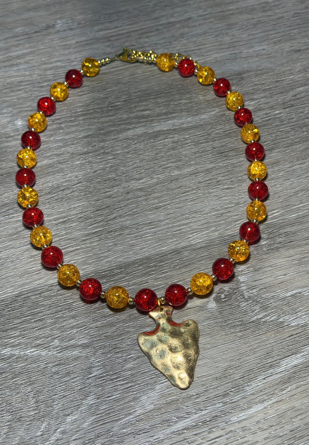 A beaded necklace with alternating translucent red and yellow faceted beads, featuring a hammered gold-tone arrowhead pendant, displayed on a gray wood-textured surface.