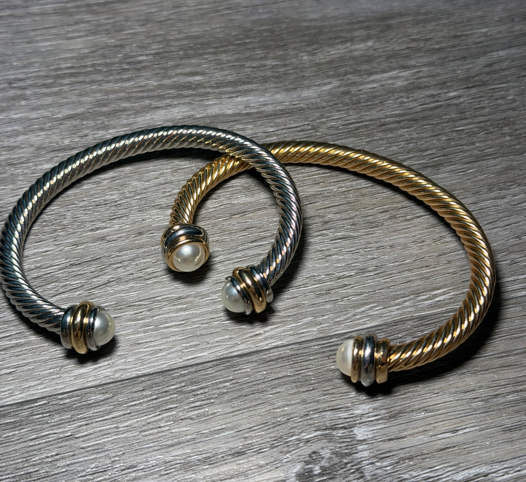 Two twisted cable-style cuff bracelets, one silver-toned and one gold-toned, each with a small white bead at the open ends, displayed on a gray wood surface.