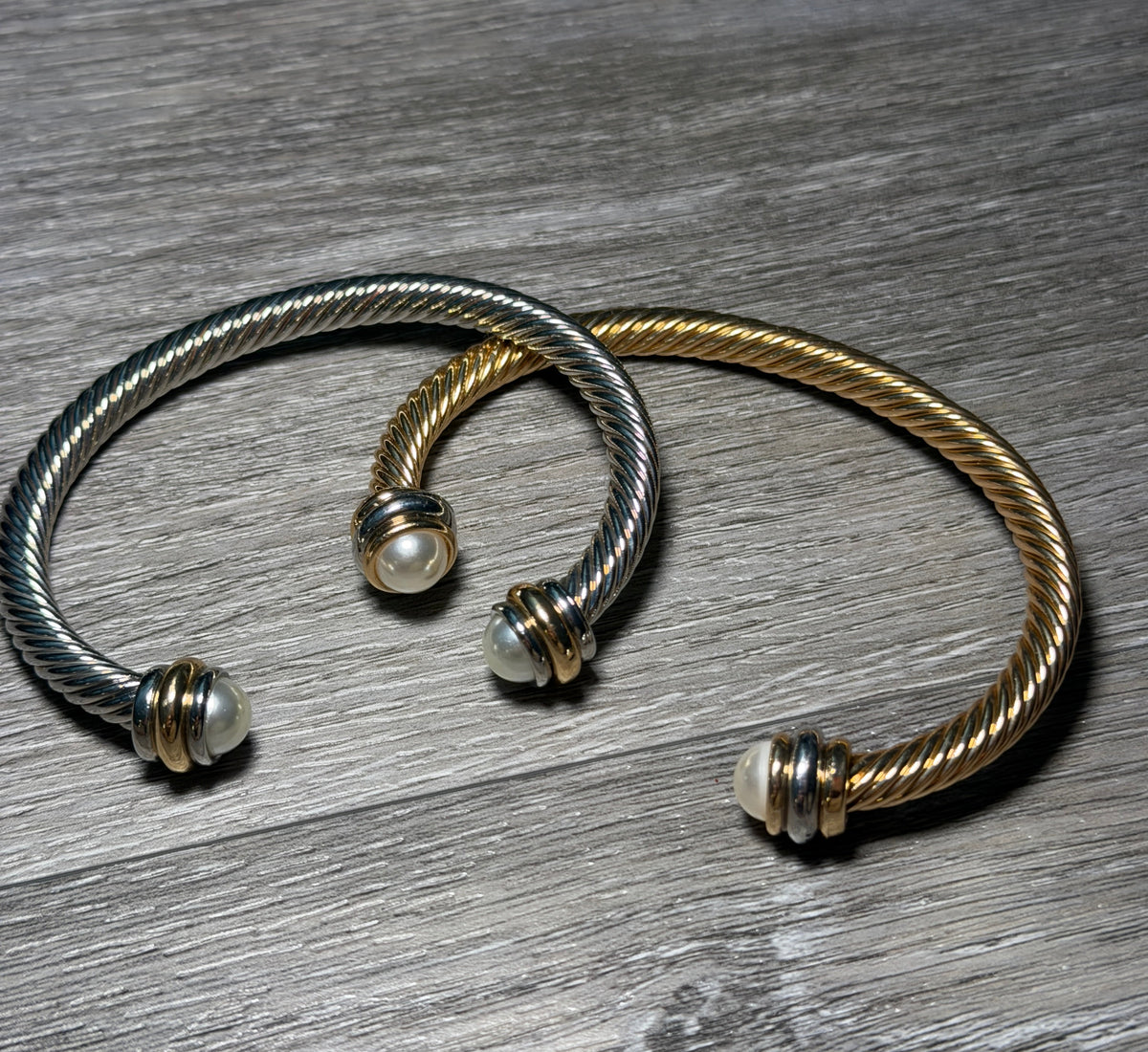 Two twisted cable-style cuff bracelets, one silver-toned and one gold-toned, each with a small white bead at the open ends, displayed on a gray wood surface.