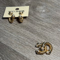 A pair of gold-toned twisted hoop earrings with small pearl accents, shown on a cream display card labeled 'Paco DESIGNS' and another pair lying on a gray wood-grain surface.