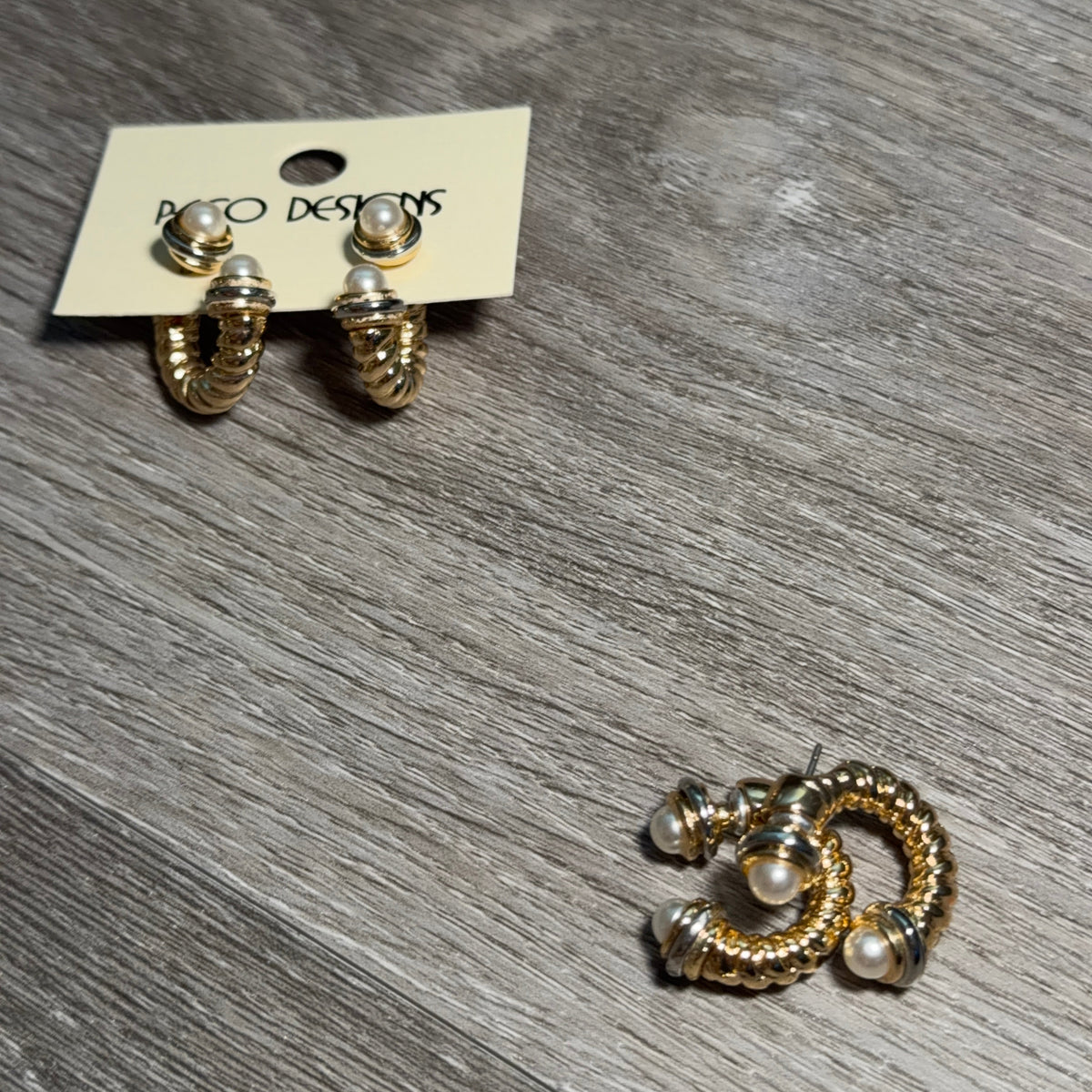 A pair of gold-toned twisted hoop earrings with small pearl accents, shown on a cream display card labeled 'Paco DESIGNS' and another pair lying on a gray wood-grain surface.