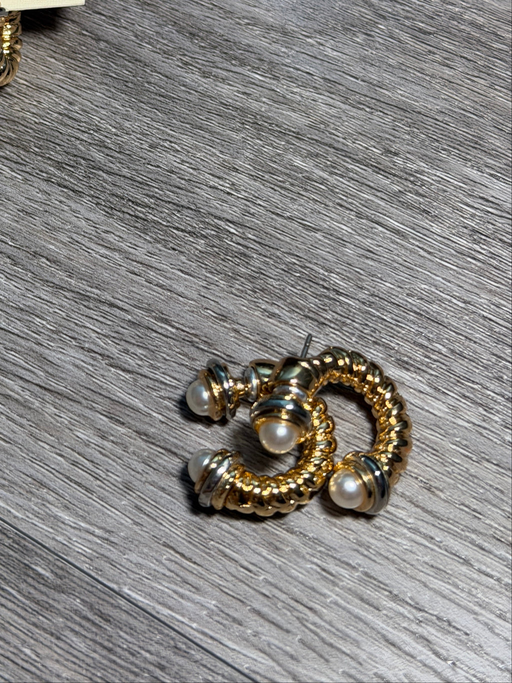 A pair of gold and silver hoop earrings with small pearl accents, shown on a cream display card and another earring placed separately on a gray wood-textured surface. The card is branded 'PACO Designs'.