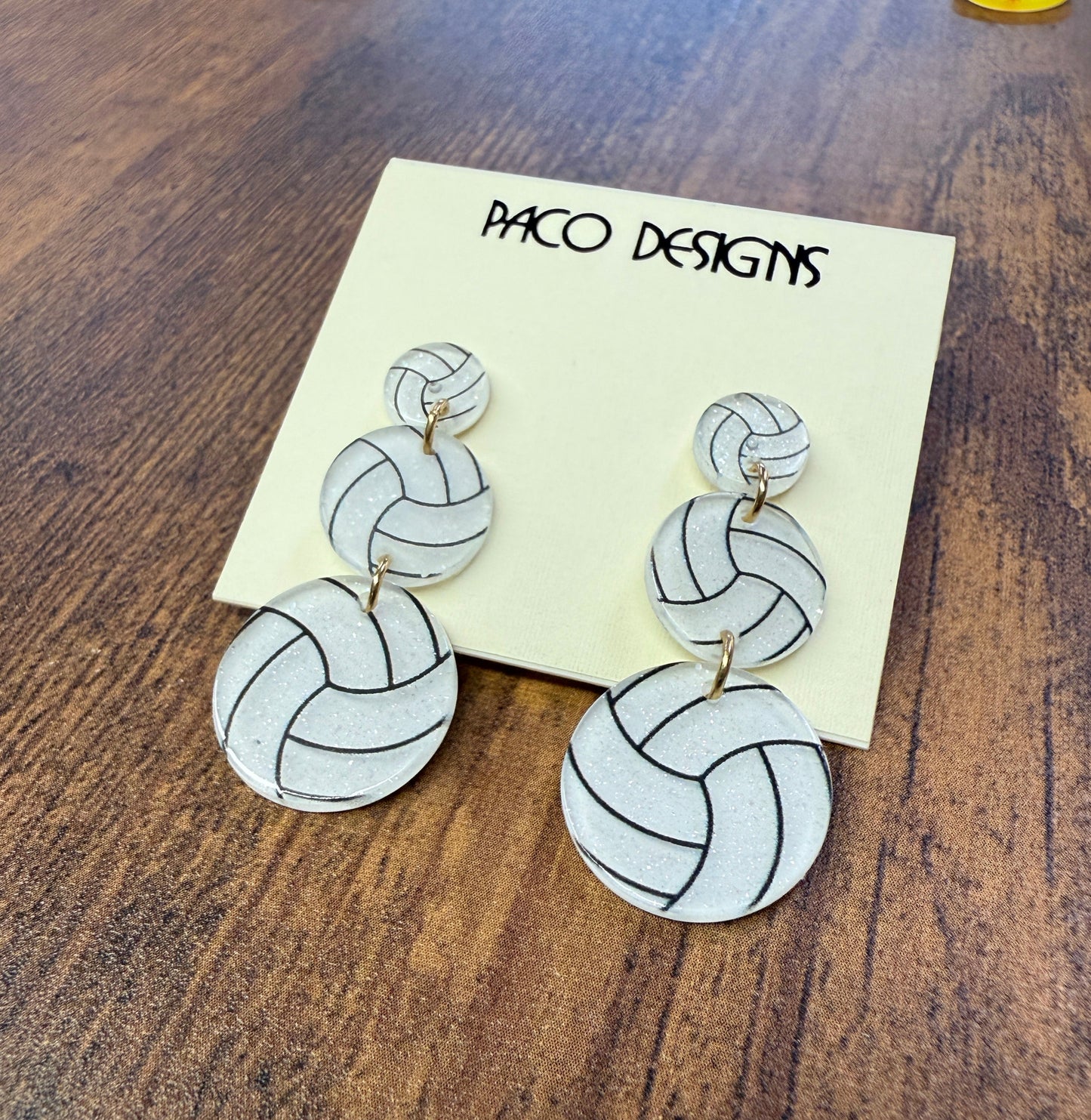 Volleyball-patterned earrings on a card with 'Paco Designs' branding on a wooden surface.