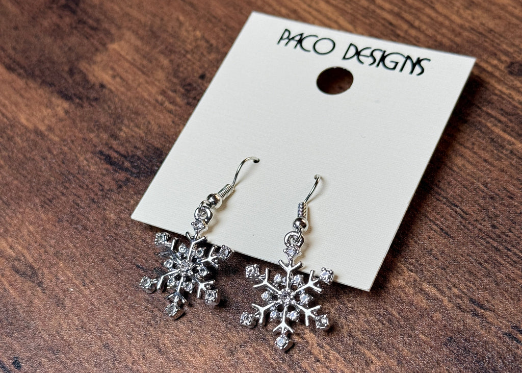 Snowflake earrings on a wooden surface with a 'Paco Designs' tag.