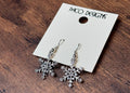 Snowflake earrings on a wooden surface with a 'Paco Designs' tag.