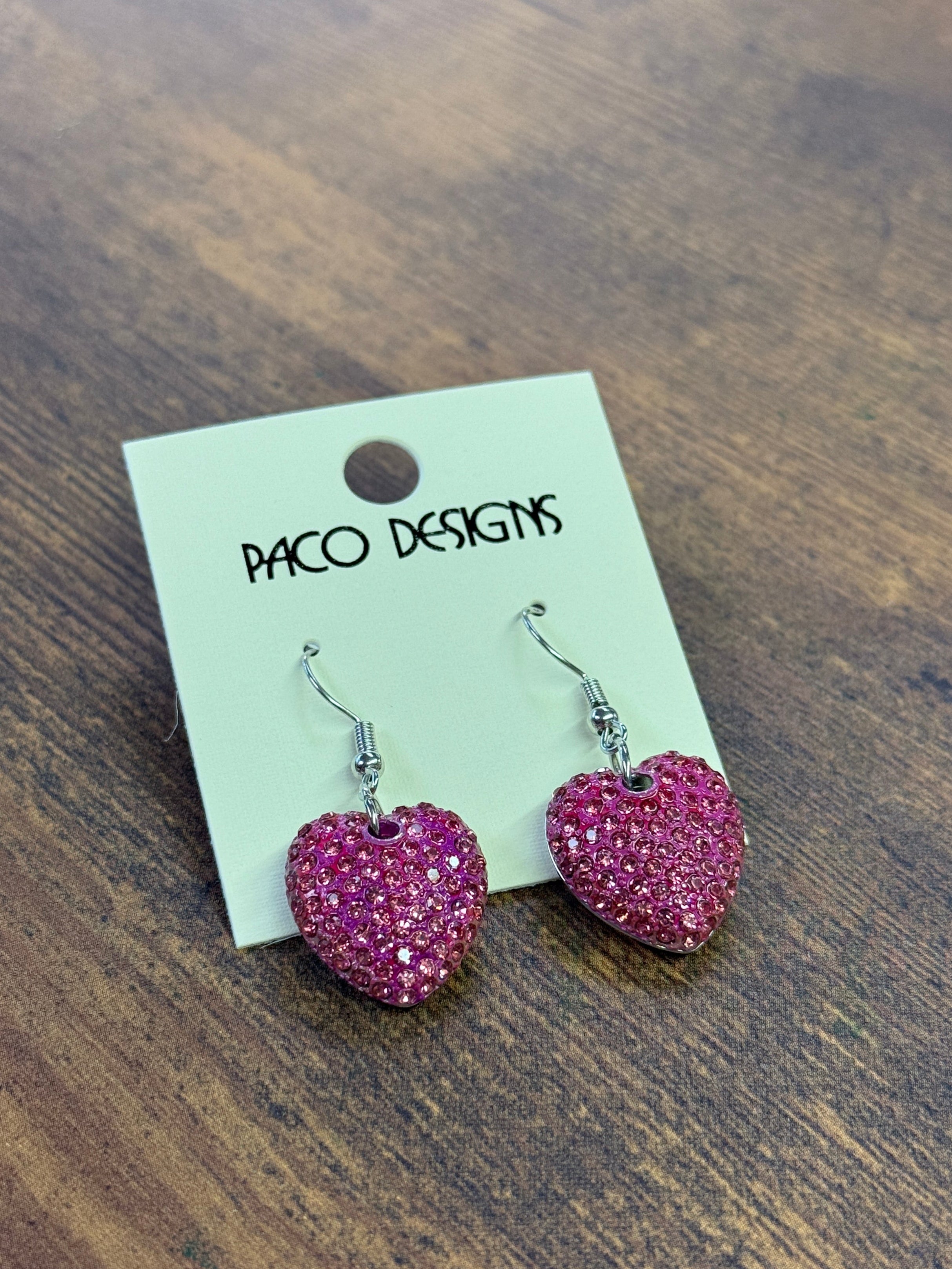 Pink heart-shaped earrings on a Paco Designs card on a wooden surface