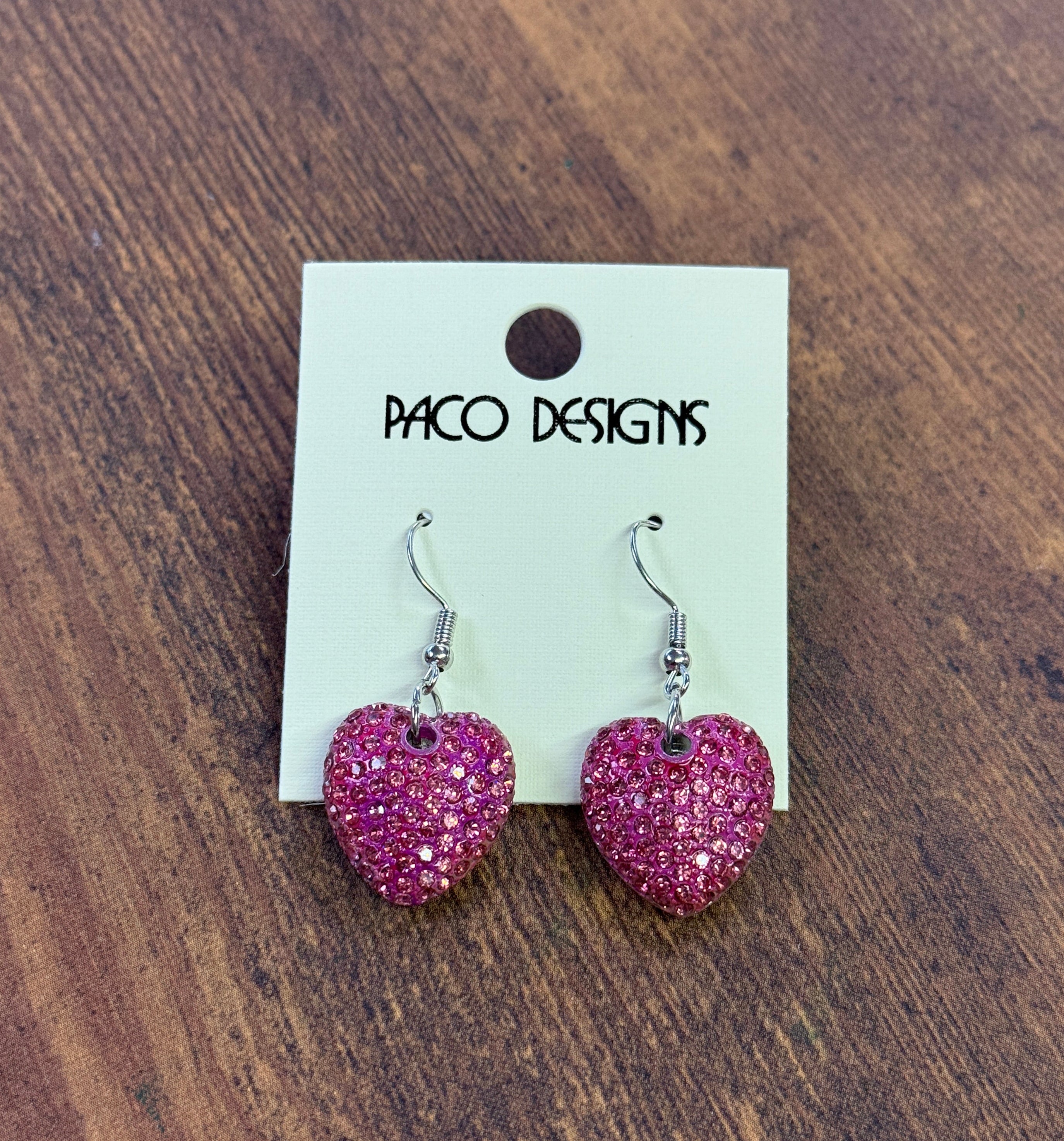 Pink heart-shaped earrings on a Paco Designs card against a wooden background