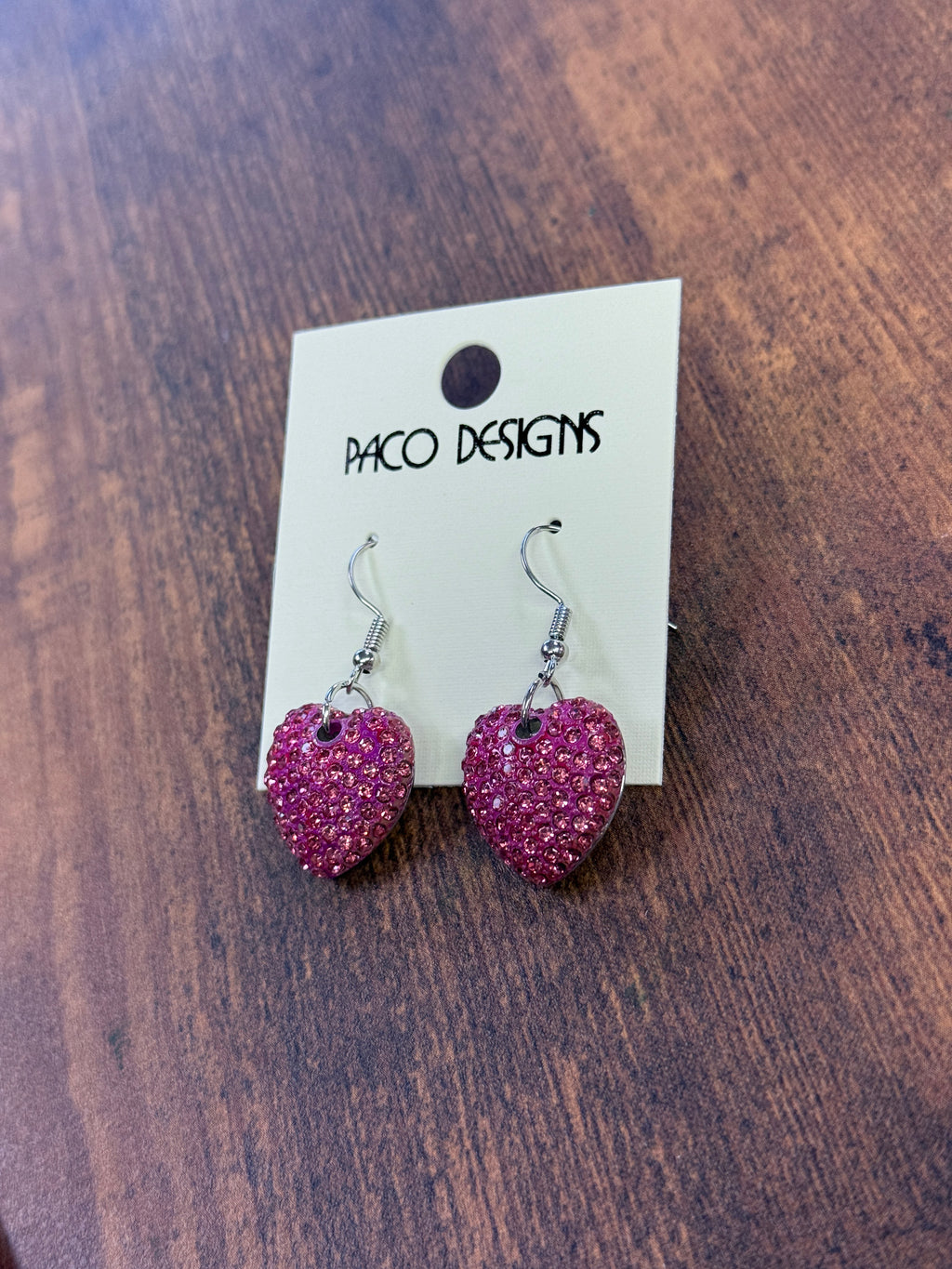 Pink heart-shaped earrings on a 'Paco Designs' card against a wooden background